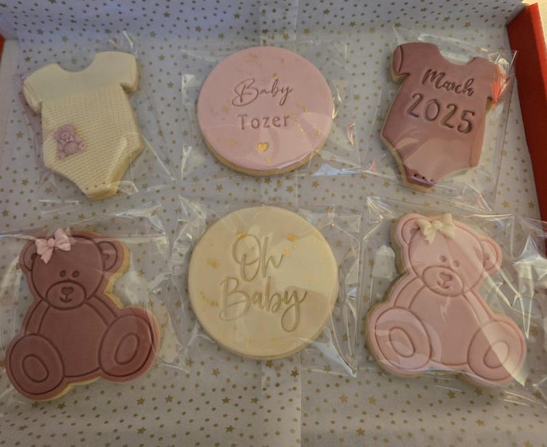 May include: Six decorated sugar cookies in a gift box. The cookies are pink and white and feature a baby onesie, a teddy bear, and the text "Baby Tozer", "Oh Baby", and "March 2025".
