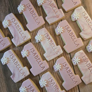 May include: A collection of number one cookies, each decorated with the name "ISABELLA" and a small white flower. The cookies have a pink frosting with a marble effect, and some have a solid pink color.