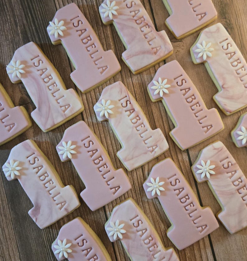 May include: A collection of number one cookies, each decorated with the name "ISABELLA" and a small white flower. The cookies have a pink frosting with a marble effect, and some have a solid pink color.