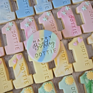 May include: An assortment of decorated sugar cookies. The cookies are in the shape of the number one, with pastel frosting in various colors. Each cookie is decorated with the name "DOTTIE". A round cookie in the center says "Happy Birthday DOTTIE".