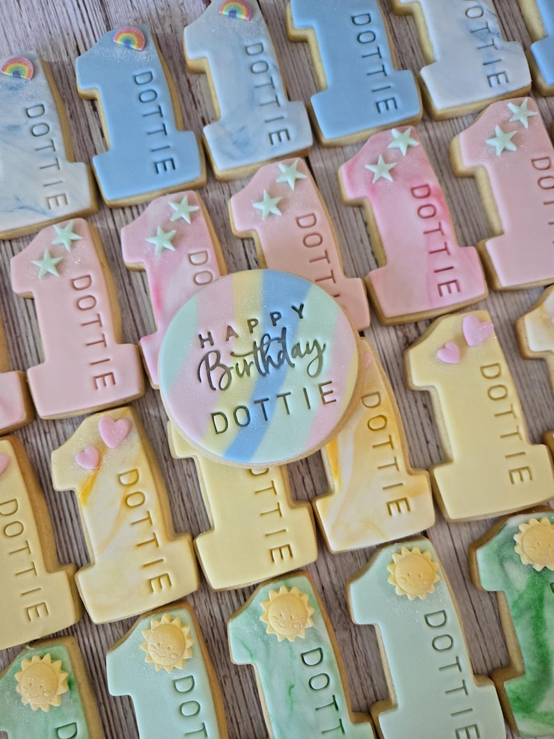 May include: An assortment of decorated sugar cookies. The cookies are in the shape of the number one, with pastel frosting in various colors. Each cookie is decorated with the name "DOTTIE". A round cookie in the center says "Happy Birthday DOTTIE".