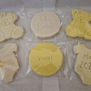 May include: Six decorated sugar cookies in individual clear plastic bags. The cookies are white and yellow and feature a baby onesie, a teddy bear, and the text "Oh Baby", "Violet", and "May 2024".