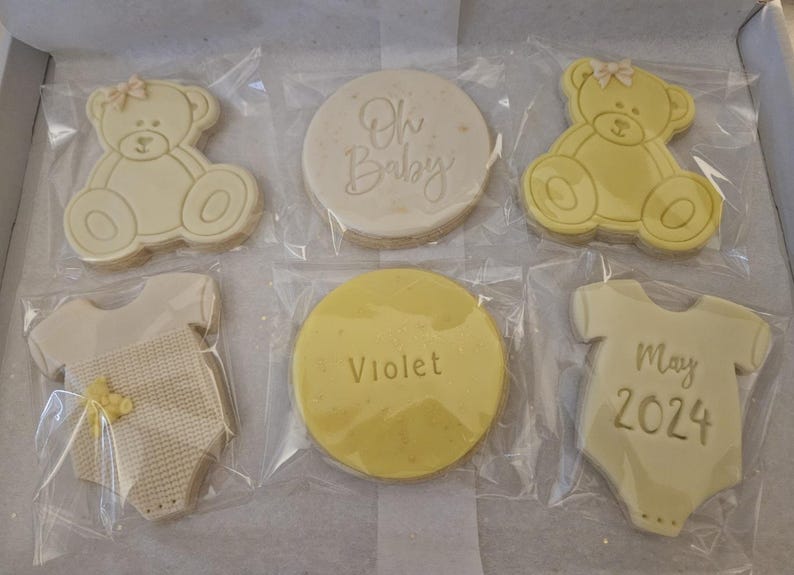 May include: Six decorated sugar cookies in individual clear plastic bags. The cookies are white and yellow and feature a baby onesie, a teddy bear, and the text "Oh Baby", "Violet", and "May 2024".
