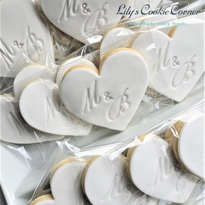 May include: Heart-shaped cookies with white icing and the initials "M & B" are displayed in clear plastic packaging. The cookies are arranged on a white tray. The logo "Lily's Cookie Corner" is visible in the top right corner.