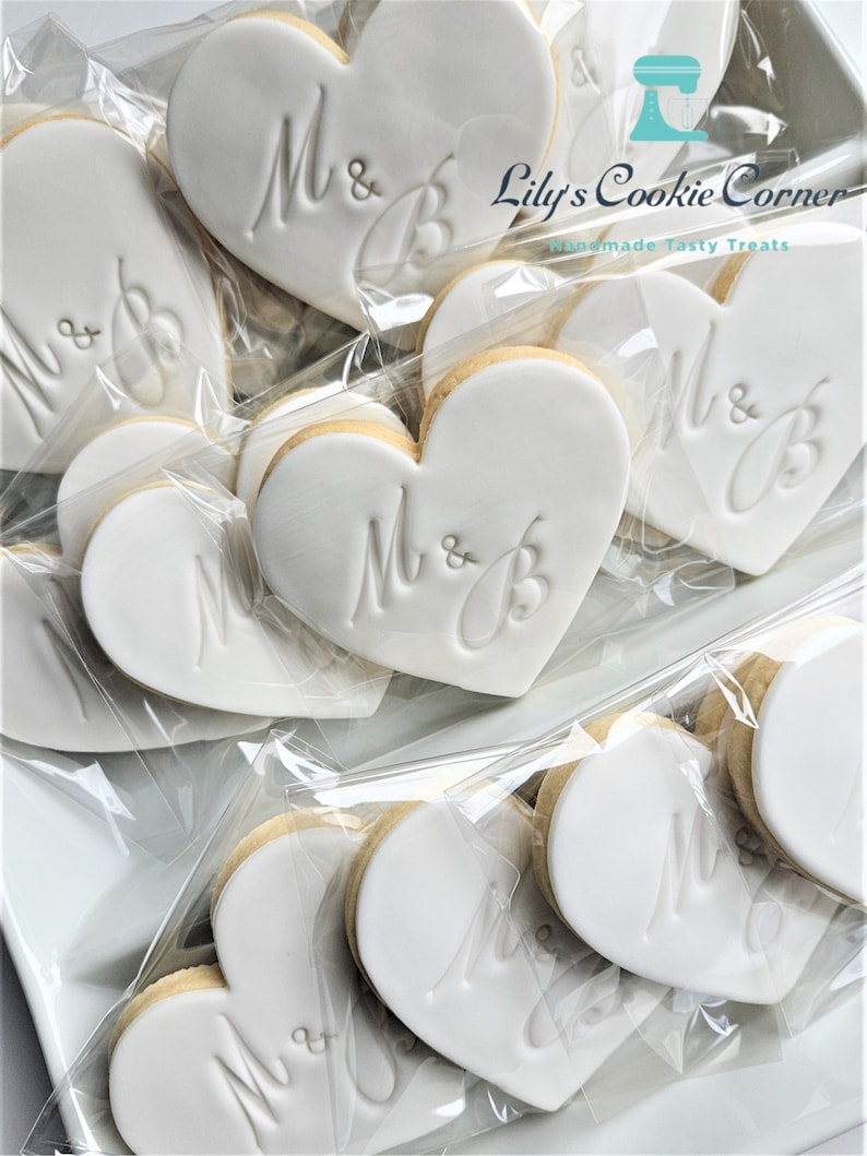 May include: Heart-shaped cookies with white icing and the initials "M & B" are displayed in clear plastic packaging. The cookies are arranged on a white tray. The logo "Lily's Cookie Corner" is visible in the top right corner.