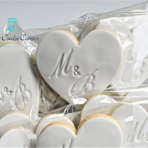 May include: White heart-shaped cookies with the initials "M & B" embossed on them, packaged in clear cellophane. The cookies have a light-colored border and are arranged in a box. The text "Lily's Cookie Corner" is visible in the upper left corner.