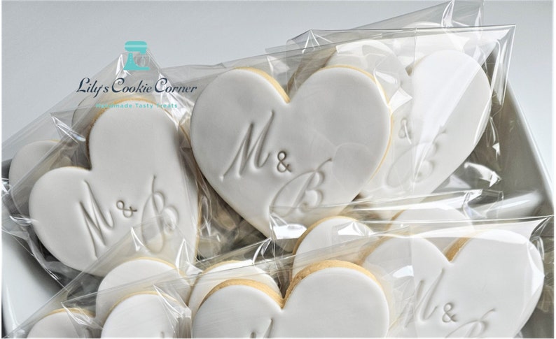 May include: White heart-shaped cookies with the initials "M & B" embossed on them, packaged in clear cellophane. The cookies have a light-colored border and are arranged in a box. The text "Lily's Cookie Corner" is visible in the upper left corner.