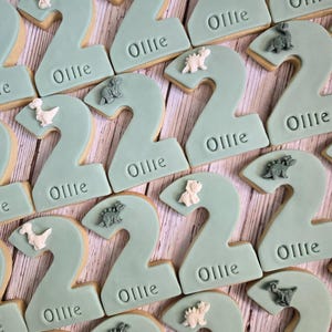 May include: A collection of number two cookies with a light teal frosting. Each cookie has the name "Ollie" imprinted on it and is decorated with small dinosaur figurines in various colors.