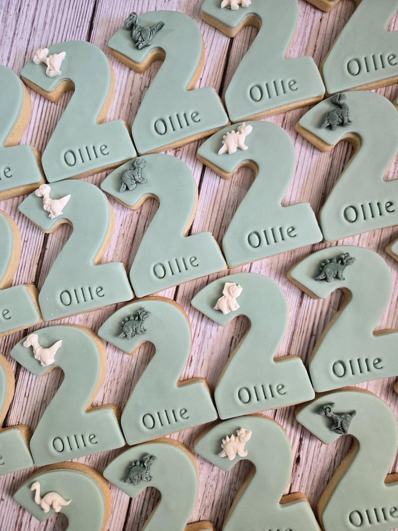 May include: A collection of number two cookies with a light teal frosting. Each cookie has the name "Ollie" imprinted on it and is decorated with small dinosaur figurines in various colors.