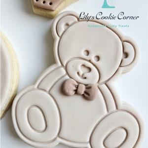 May include: A light brown sugar cookie decorated with a light brown teddy bear design. The teddy bear has a brown bow tie and is outlined in a darker brown. The cookie is a handmade tasty treat from Lily's Cookie Corner.