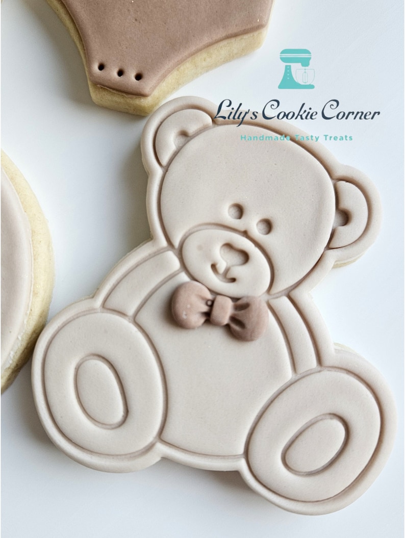 May include: A light brown sugar cookie decorated with a light brown teddy bear design. The teddy bear has a brown bow tie and is outlined in a darker brown. The cookie is a handmade tasty treat from Lily's Cookie Corner.