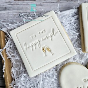 Pode incluir: Um biscoito quadrado branco com o texto em relevo "TO THE happy couple" e duas ta&ccedil;as de champanhe. O biscoito est&aacute; numa caixa com papel branco. Um presente de casamento ou noivado.