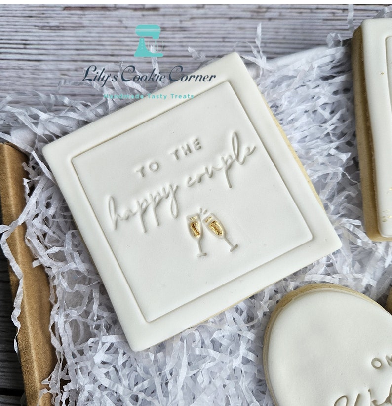 Pode incluir: Um biscoito quadrado branco com o texto em relevo "TO THE happy couple" e duas ta&ccedil;as de champanhe. O biscoito est&aacute; numa caixa com papel branco. Um presente de casamento ou noivado.