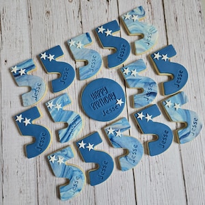 May include: An assortment of birthday cookies arranged on a wooden surface. The cookies are primarily blue and white, shaped like the number 5 and a circle. The number 5 cookies have white star decorations and the name "Jesse". The circular cookie says "Happy Birthday Jesse".
