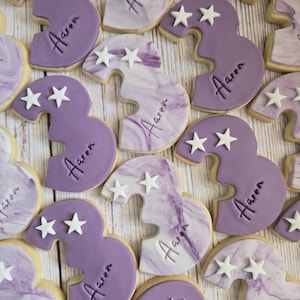 May include: Overhead shot of several number three cookies. The cookies are decorated with purple frosting and white star sprinkles. Each cookie has the name "Aaron" written on it.