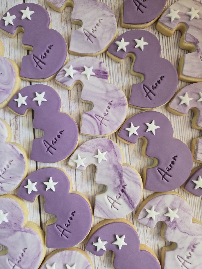 May include: Overhead shot of several number three cookies. The cookies are decorated with purple frosting and white star sprinkles. Each cookie has the name "Aaron" written on it.