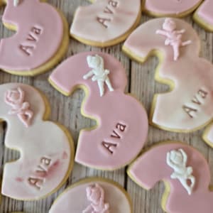 May include: Pink and white frosted number three cookies with the name "Ava" and small ballerina figures. The cookies have a light pink frosting and a golden-brown edge. The ballerina figures are white and pink.