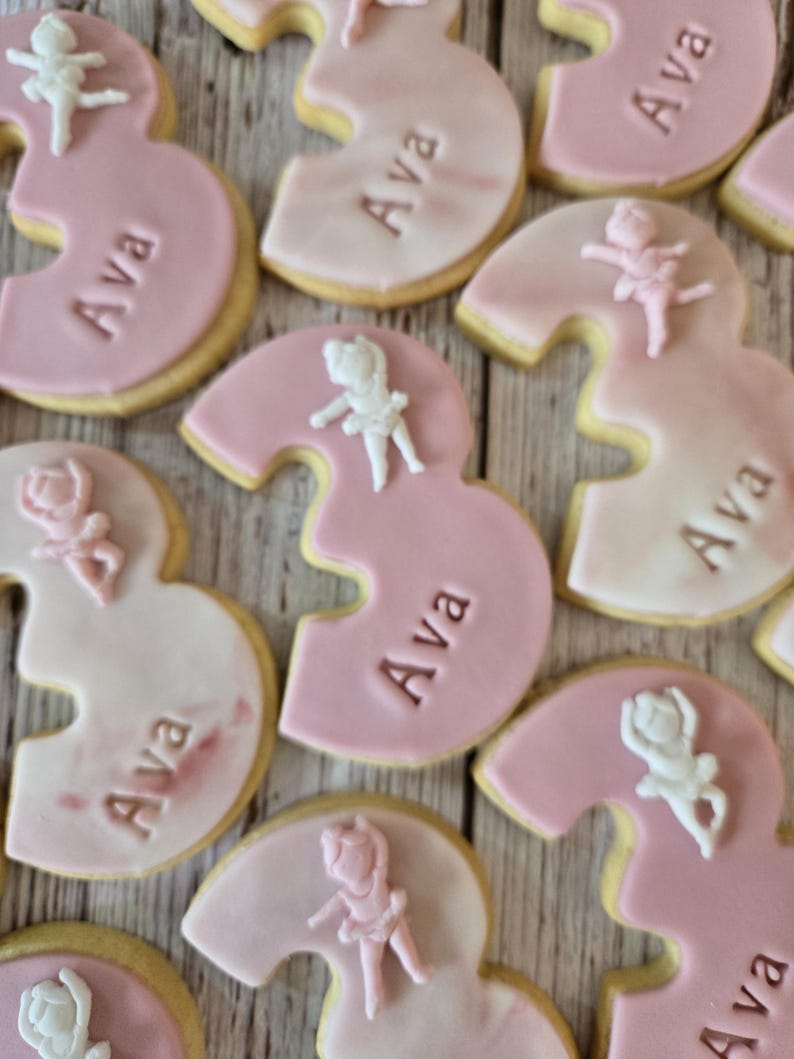 May include: Pink and white frosted number three cookies with the name "Ava" and small ballerina figures. The cookies have a light pink frosting and a golden-brown edge. The ballerina figures are white and pink.