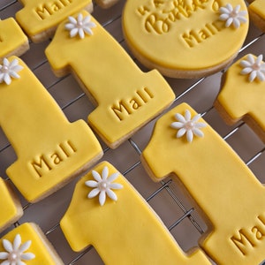 May include: Yellow frosted cookies, some shaped as the number one, and others round, with the name "Mall" and "Happy Birthday Mall" embossed. Each number one cookie is decorated with a white flower with a pearl center. The cookies are arranged on a metal cooling rack.