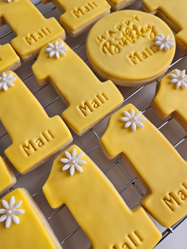 May include: Yellow frosted cookies, some shaped as the number one, and others round, with the name "Mall" and "Happy Birthday Mall" embossed. Each number one cookie is decorated with a white flower with a pearl center. The cookies are arranged on a metal cooling rack.