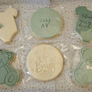 May include: A set of six decorated sugar cookies in a light blue and white color scheme. The cookies feature a baby onesie, a teddy bear, and a round cookie with the text "Oh Baby" and "Baby AV" in gold lettering. The cookies are packaged in clear plastic bags.
