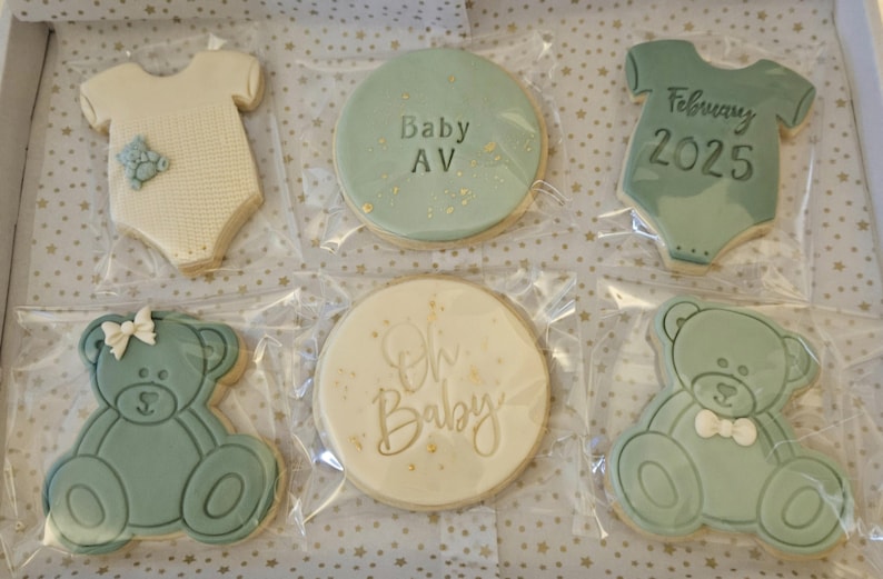 May include: A set of six decorated sugar cookies in a light blue and white color scheme. The cookies feature a baby onesie, a teddy bear, and a round cookie with the text "Oh Baby" and "Baby AV" in gold lettering. The cookies are packaged in clear plastic bags.
