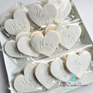 May include: Heart-shaped cookies with white icing, each individually wrapped in clear plastic. The cookies are decorated with the letters "M & B" in an elegant script. The cookies are arranged on a white rectangular tray.
