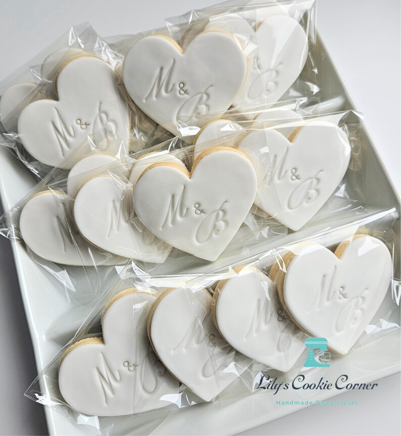 May include: Heart-shaped cookies with white icing, each individually wrapped in clear plastic. The cookies are decorated with the letters "M & B" in an elegant script. The cookies are arranged on a white rectangular tray.