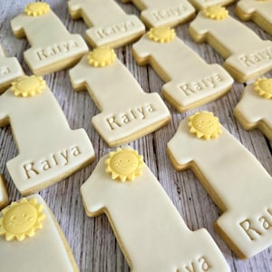 May include: Close-up of several number one cookies with white icing and a yellow sun decoration. The name "Kalya" is written on each cookie. The cookies are arranged on a light-colored wooden surface.