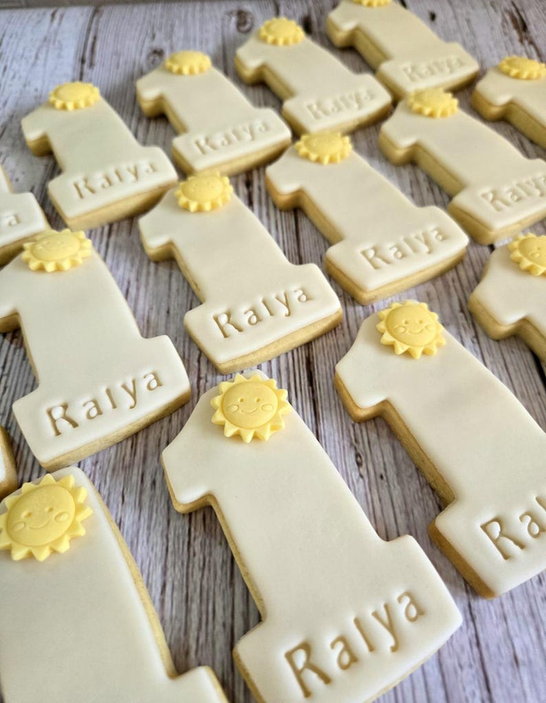 May include: Close-up of several number one cookies with white icing and a yellow sun decoration. The name "Kalya" is written on each cookie. The cookies are arranged on a light-colored wooden surface.