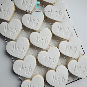 May include: Heart-shaped cookies with white icing and the initials "M & B" are arranged on a cooling rack. The cookies have a light brown edge and are likely for a wedding or engagement celebration. "Lily's Cookie Corner" is in the image.