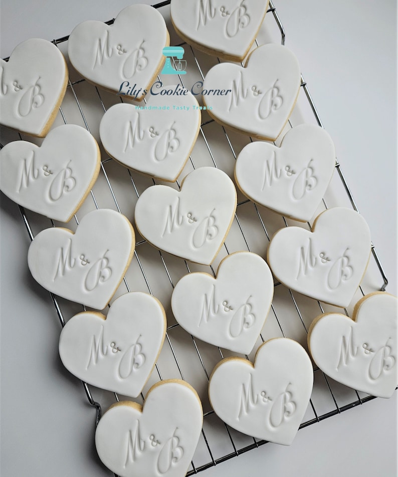 May include: Heart-shaped cookies with white icing and the initials "M & B" are arranged on a cooling rack. The cookies have a light brown edge and are likely for a wedding or engagement celebration. "Lily's Cookie Corner" is in the image.