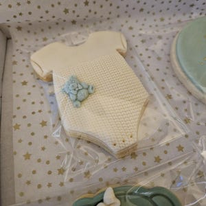 May include: A white cookie shaped like a baby onesie with a blue knitted texture and a blue teddy bear applique. The cookie is on a white background with gold stars.