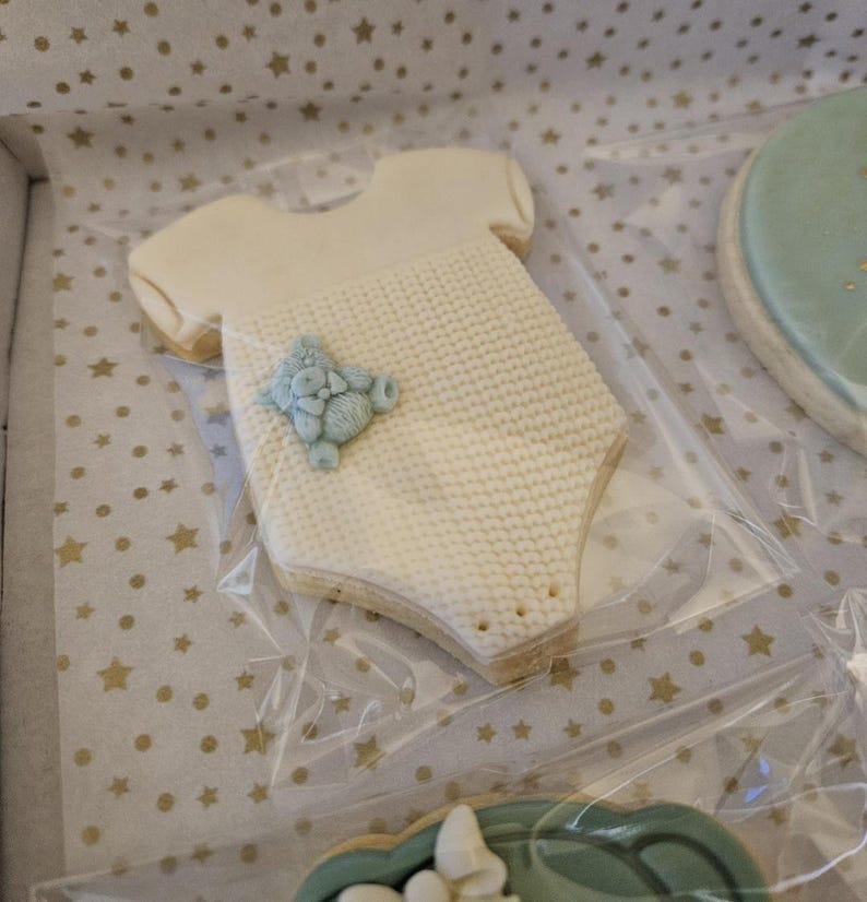 May include: A white cookie shaped like a baby onesie with a blue knitted texture and a blue teddy bear applique. The cookie is on a white background with gold stars.