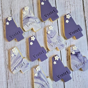 May include: A collection of custom-decorated cookies in the shape of the number four. The cookies are primarily purple with white floral accents and the name "Violet" written on them. Some cookies have a marbled design.