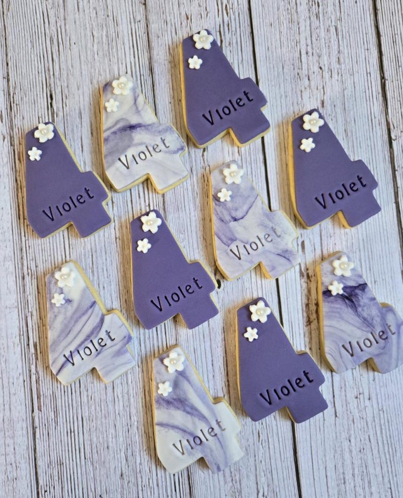 May include: A collection of custom-decorated cookies in the shape of the number four. The cookies are primarily purple with white floral accents and the name "Violet" written on them. Some cookies have a marbled design.