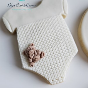 May include: White sugar cookie decorated to look like a baby onesie with a knitted texture and a small brown teddy bear on the front. The cookie is decorated with the text "Lily's Cookie Corner Handmade Tasty Treats".