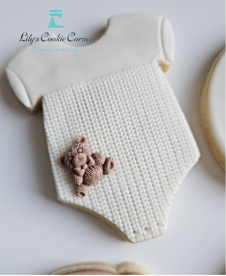May include: White sugar cookie decorated to look like a baby onesie with a knitted texture and a small brown teddy bear on the front. The cookie is decorated with the text "Lily's Cookie Corner Handmade Tasty Treats".