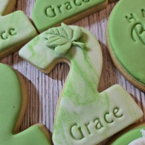 May include: Close-up of decorated sugar cookies. The cookies are in the shape of the number two, with green and white marbled icing. The name "Grace" is imprinted on the cookies. A leaf-shaped decoration is on top of one cookie.