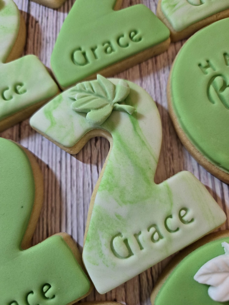 May include: Close-up of decorated sugar cookies. The cookies are in the shape of the number two, with green and white marbled icing. The name "Grace" is imprinted on the cookies. A leaf-shaped decoration is on top of one cookie.
