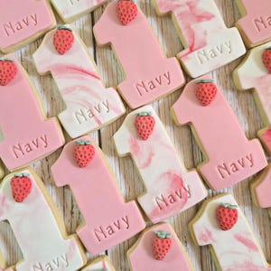May include: A collection of number one cookies in various shades of pink and white, decorated with a strawberry on top. The word "Navy" is imprinted on each cookie. The cookies are arranged on a light wooden surface.