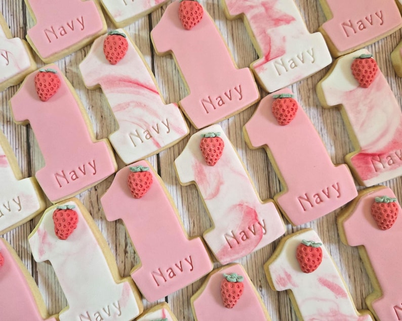 May include: A collection of number one cookies in various shades of pink and white, decorated with a strawberry on top. The word "Navy" is imprinted on each cookie. The cookies are arranged on a light wooden surface.