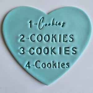 May include: A light blue heart-shaped cookie cutter with the words "1-Cookies", "2-COOKIES", "3-COOKIES", and "4-Cookies" embossed on the surface. The cookie cutter is made of a smooth material and is designed for baking.