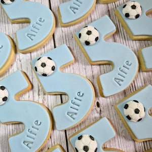May include: Light blue number five cookies with the name "Alfie" and a soccer ball decoration. The cookies are arranged on a light wood grain surface. The cookies are likely for a birthday celebration.