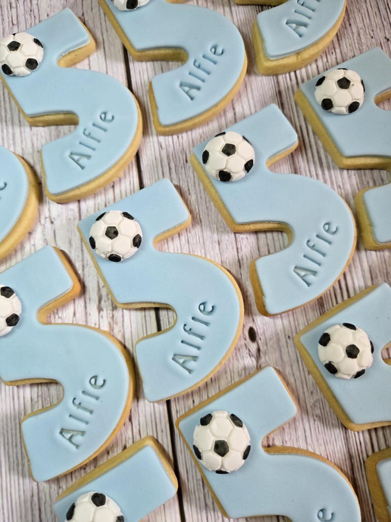 May include: Light blue number five cookies with the name "Alfie" and a soccer ball decoration. The cookies are arranged on a light wood grain surface. The cookies are likely for a birthday celebration.
