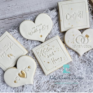 Pode incluir: Uma cole&ccedil;&atilde;o de biscoitos decorados numa bandeja de madeira, perfeitos para um casamento. Os biscoitos s&atilde;o brancos com detalhes dourados e v&aacute;rias mensagens, incluindo "Parab&eacute;ns!" e "No seu dia de casamento". Alguns biscoitos s&atilde;o em forma de cora&ccedil;&atilde;o, outros quadrados, com a data 12/07/2024.