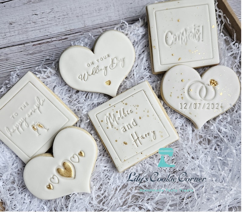 Pode incluir: Uma cole&ccedil;&atilde;o de biscoitos decorados numa bandeja de madeira, perfeitos para um casamento. Os biscoitos s&atilde;o brancos com detalhes dourados e v&aacute;rias mensagens, incluindo "Parab&eacute;ns!" e "No seu dia de casamento". Alguns biscoitos s&atilde;o em forma de cora&ccedil;&atilde;o, outros quadrados, com a data 12/07/2024.
