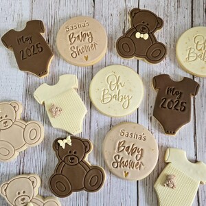 May include: A collection of nine decorated sugar cookies for a baby shower. The cookies feature a variety of designs, including teddy bears, onesies, and text that says "Sasha's Baby Shower", "Oh Baby", and "May 2025".
