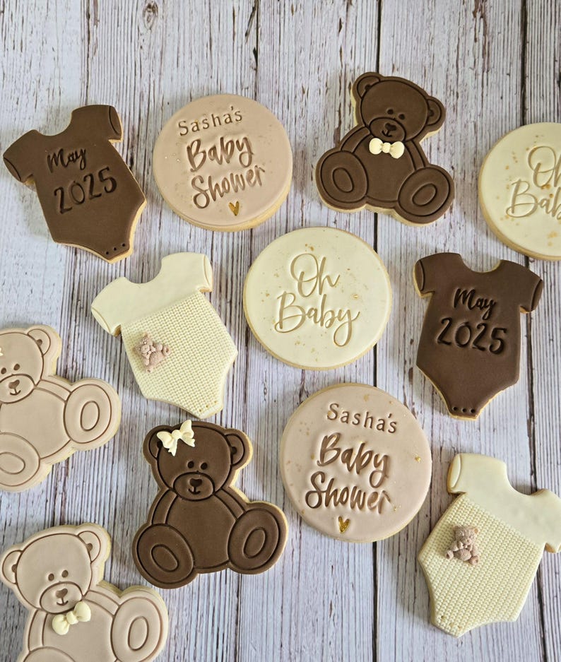 May include: A collection of nine decorated sugar cookies for a baby shower. The cookies feature a variety of designs, including teddy bears, onesies, and text that says "Sasha's Baby Shower", "Oh Baby", and "May 2025".