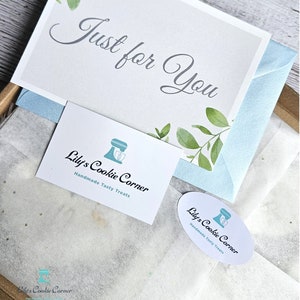 Pode incluir: Uma caixa de presente com um cart&atilde;o que diz "Just for You" em uma escrita elegante, adornado com ilustra&ccedil;&otilde;es de folhas verdes. A caixa inclui um envelope azul claro e cart&otilde;es de visita com o logotipo "Lily's Cookie Corner" e o texto "Handmade Tasty Treats."